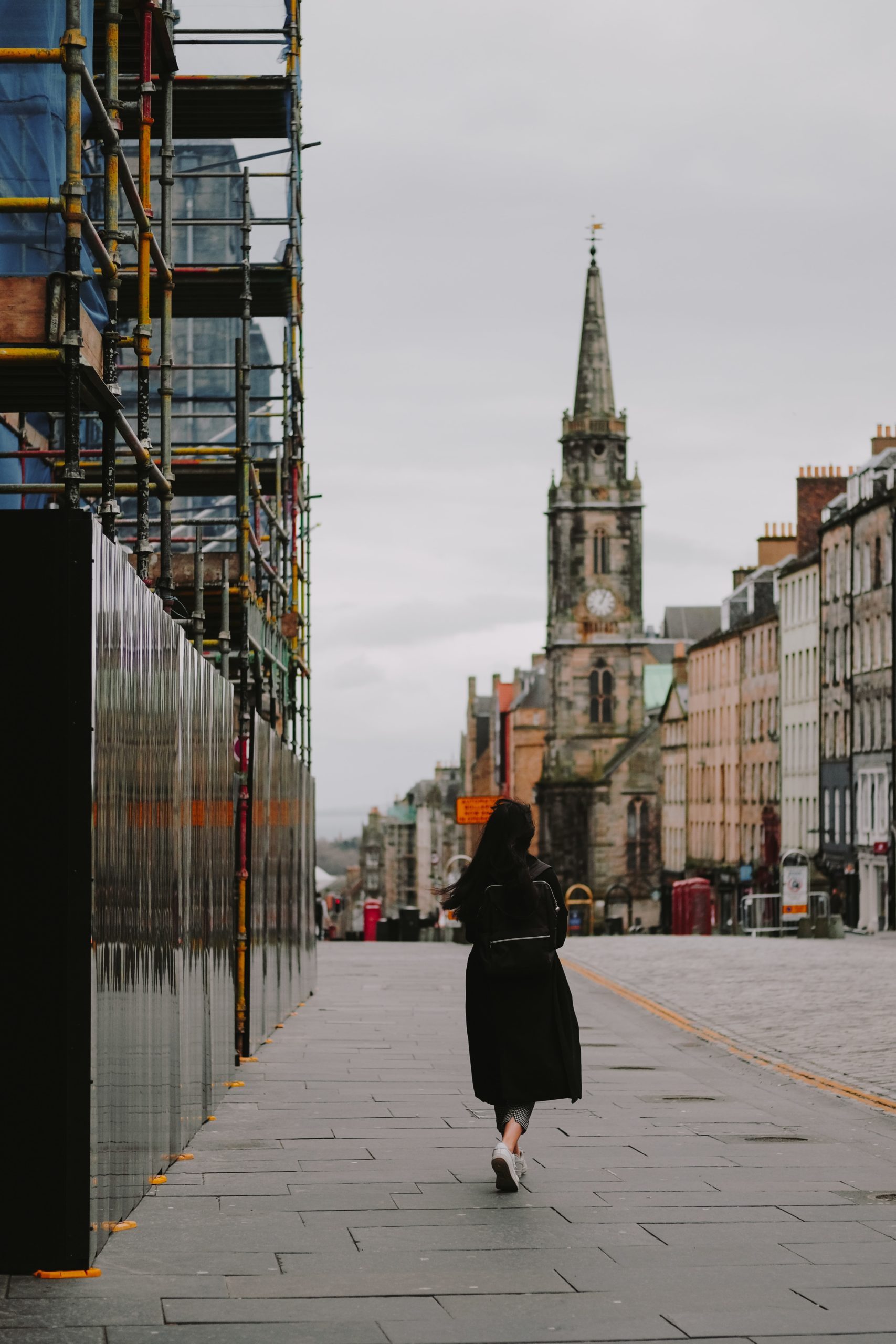 A woman in a black coat walks down a street. The sky is grey and you can see a tall building with an old, ornate roof in the background.